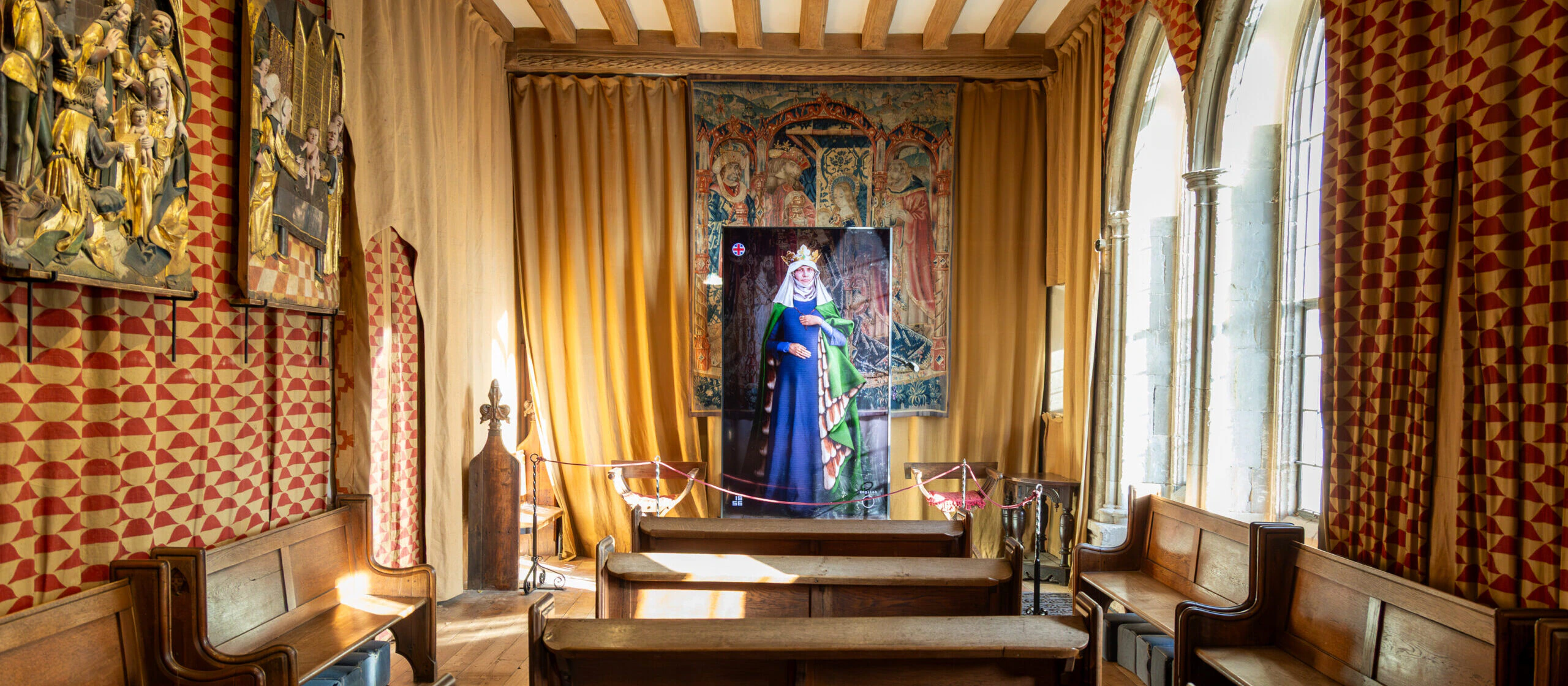 A historic chapel with wooden pews, tall arched windows, patterned walls, and a large religious painting of a crowned figure at the front, illuminated by sunlight streaming through the windows.