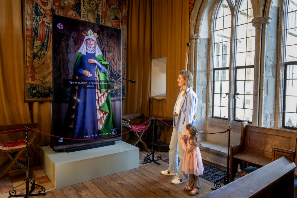 A woman and a young girl stand in front of a digital screen displaying a medieval queen in a historic, ornate room with tall windows and tapestries. The girl looks up at the screen, which has a microphone in front of it.