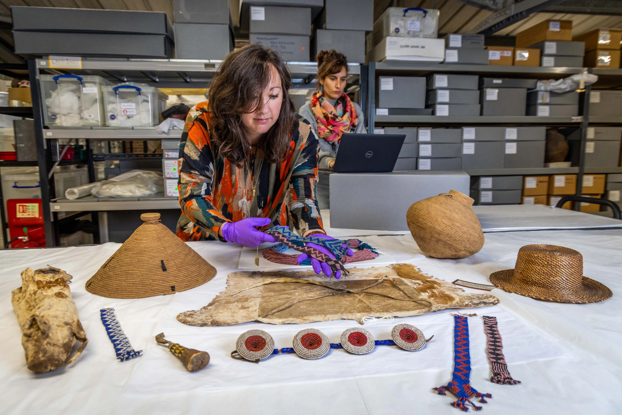 Two women work in a storage room with shelves and boxes. One woman examines colorful woven bands on a table with hats, artifacts, and animal hide, while the other uses a laptop in the background.