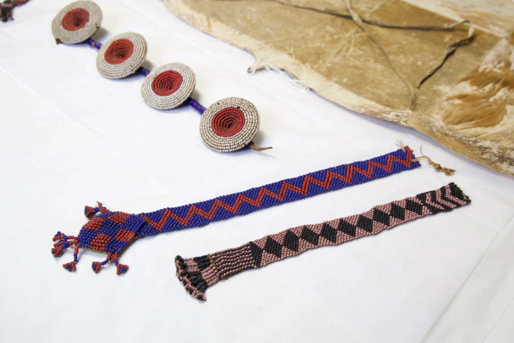 Three beaded items, including a circular patterned necklace and two beaded bracelets—one blue with red zigzags, the other pink and black—are displayed on a white cloth next to a piece of animal hide.