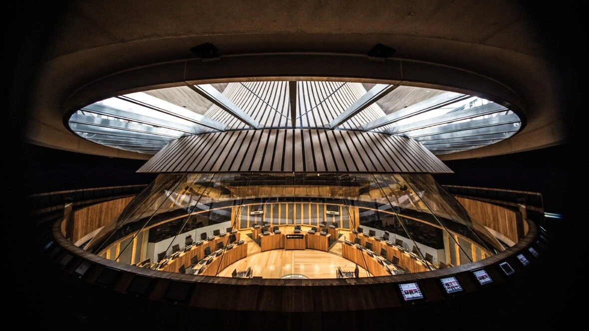 A modern, circular parliamentary chamber with tiered wooden seating, glass barriers, and a domed skylight ceiling allowing natural light to illuminate the room.