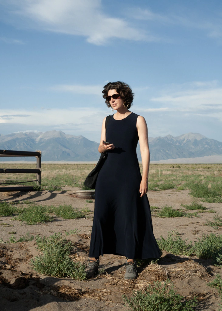 A woman in a long navy dress and sunglasses stands outdoors on a sunny day, with mountains and a partly cloudy sky in the background. She holds something in her hand and has short, dark curly hair.