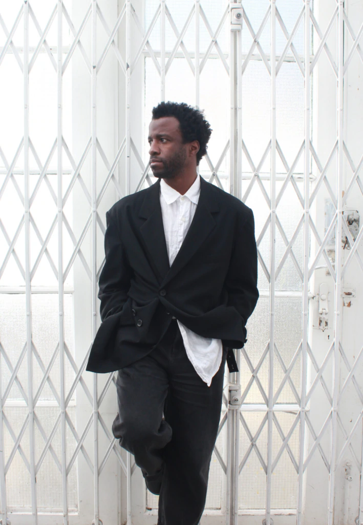 A man with short curly hair, wearing a black blazer over a white shirt and dark pants, stands in front of a white metal gate with a geometric pattern, looking to his left with his hands in his pockets.