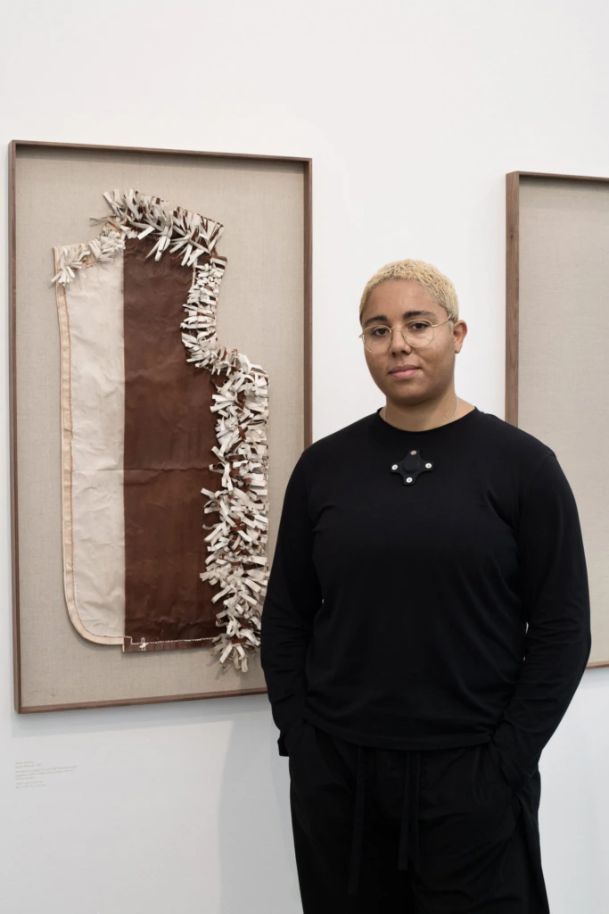 A person with short blond hair, glasses, and a black outfit stands in front of a framed textile artwork featuring brown and beige fabrics with textured, layered details.