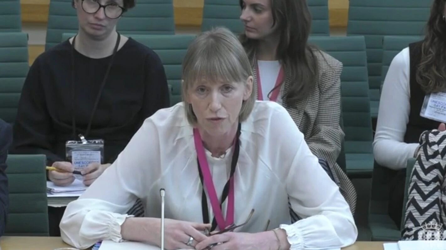 A woman with light brown hair wearing a white blouse and pink lanyard speaks at a desk with a microphone, while three women sit behind her in a formal setting with green seats.