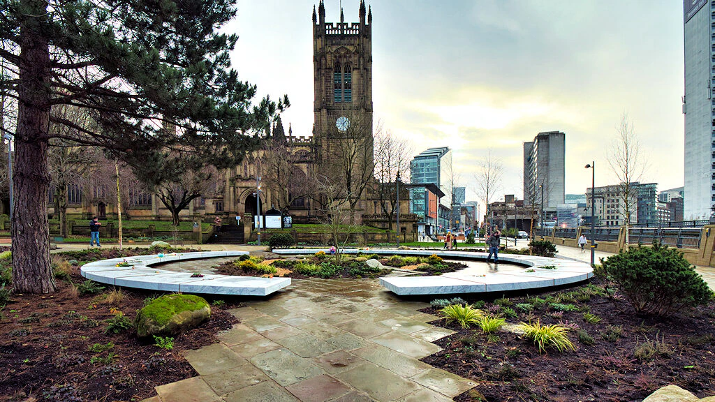 A stone path leads through landscaped gardens towards a historic church with a tall clock tower, set against a city skyline with modern buildings under a cloudy sky. People walk and enjoy the urban park.