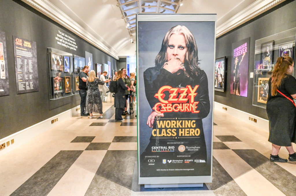 A museum exhibit hallway features a large display promoting Ozzy Osbourne: Working Class Hero. Visitors are viewing photos and memorabilia on the walls in a well-lit, modern gallery space.