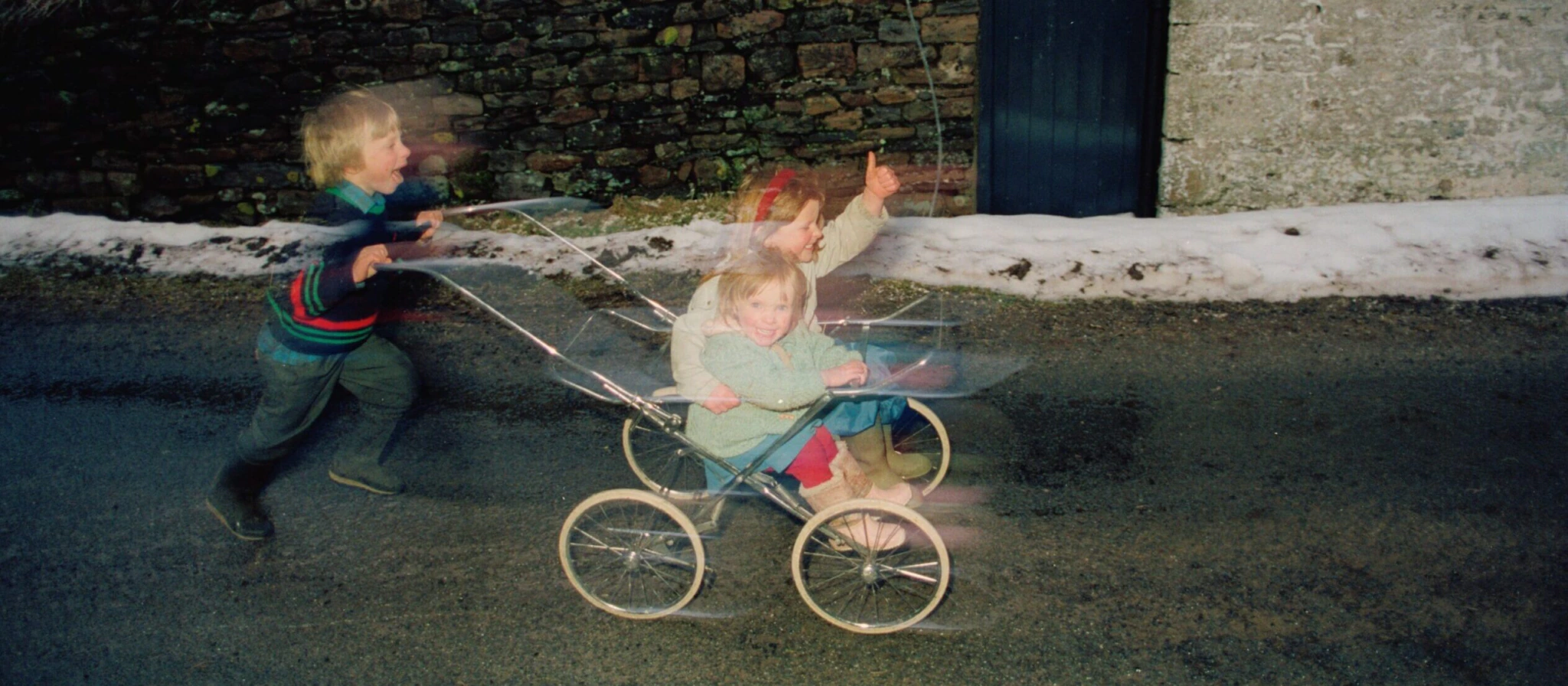 Two children sit in a vintage stroller while another child pushes them quickly down a road. The image has motion blur, with a stone wall and a Waters Meeting sign visible in the snowy background.