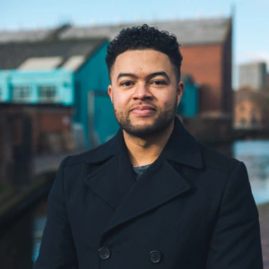 A man with short curly hair and a trimmed beard, wearing a dark coat, stands by a canal with brick buildings and a blue shed in the background on a clear day.