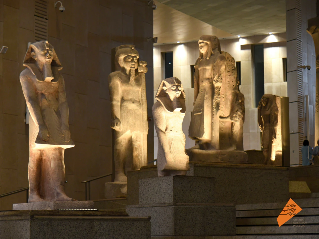 Several ancient Egyptian statues stand on display inside a museum, illuminated by warm lighting. The statues vary in size and posture, and are arranged on tiered stone platforms. An orange logo is visible in the lower right corner.