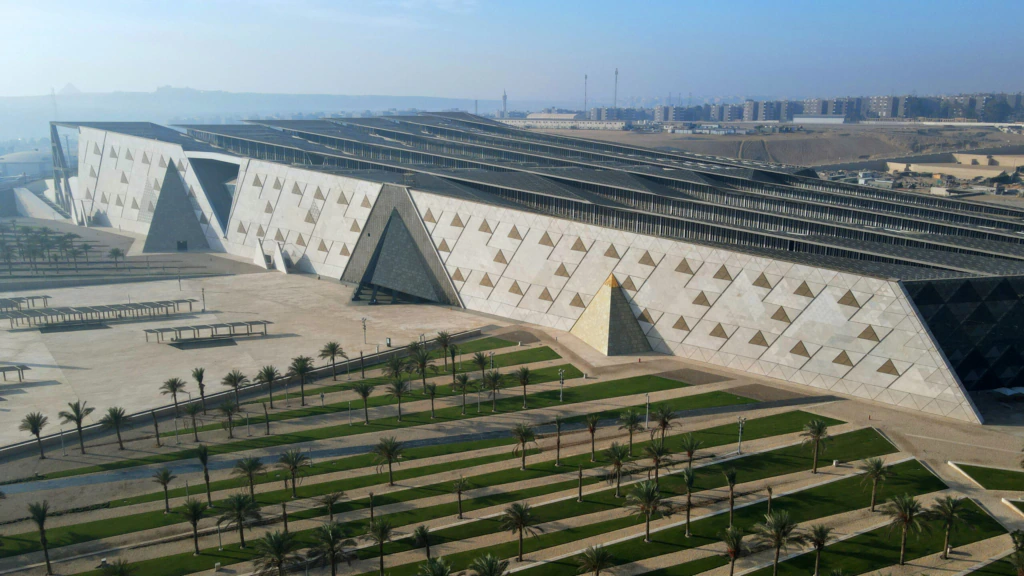Aerial view of the Grand Egyptian Museum in Giza, featuring a modern, angular facade with triangular patterns, surrounded by palm trees and landscaped green areas, with a cityscape in the background.