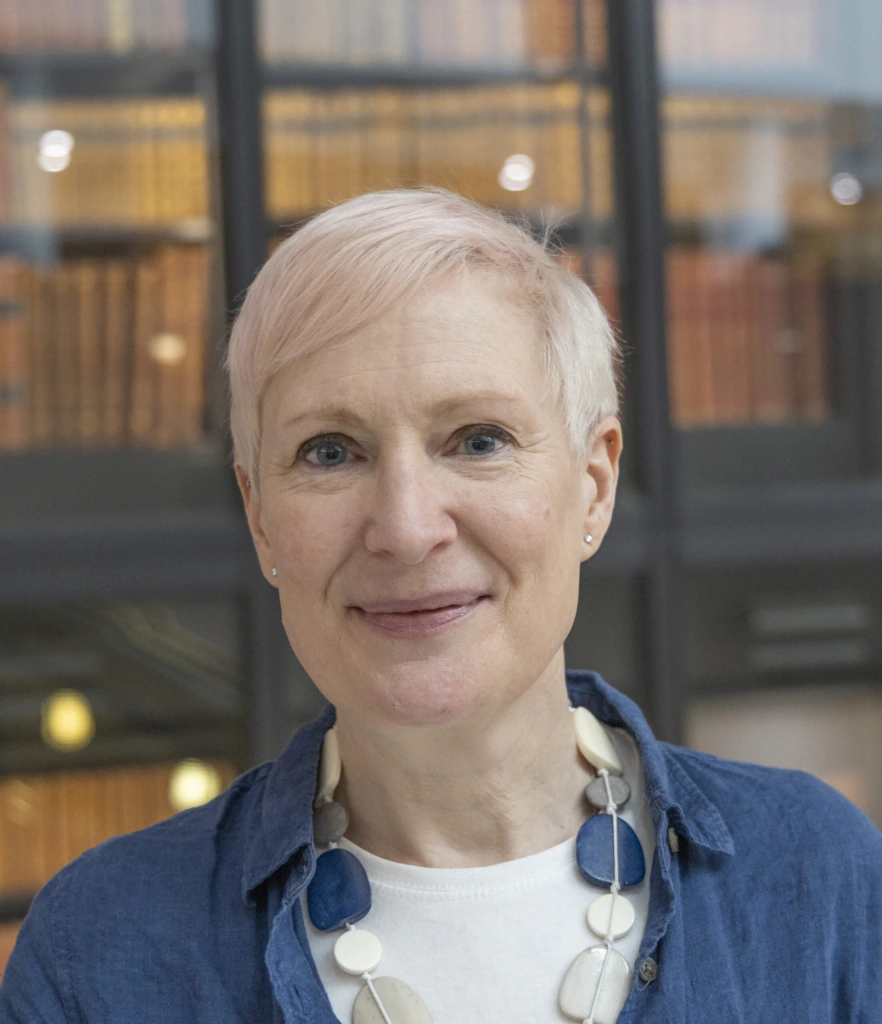 A person with short, light pink hair smiles softly at the camera. They are wearing a blue shirt, a white top, and a chunky patterned necklace, with bookshelves blurred in the background.