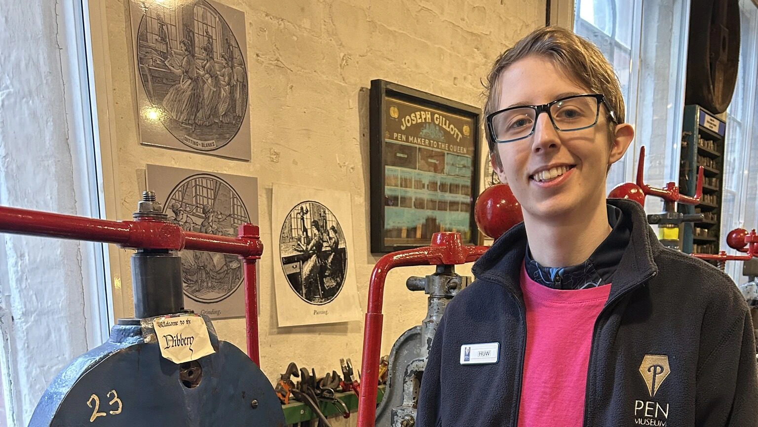 A smiling person wearing glasses and a navy fleece stands by a blue industrial machine in a workshop, with red pipes and framed pictures on the cream brick wall behind them.