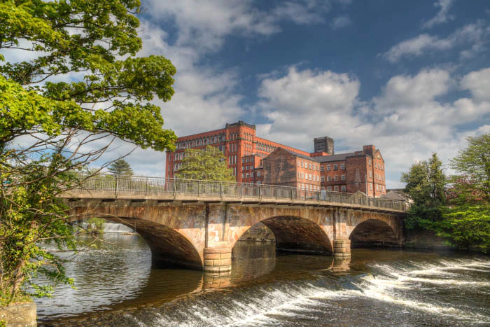 A stone bridge with three arches crosses a river with a small weir. Behind the bridge stands a large historic red brick mill building under a partly cloudy sky, surrounded by green trees.