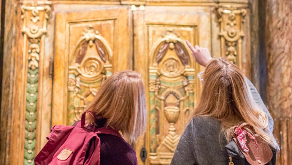 Two people with backpacks stand in front of an ornate wooden door, decorated with intricate carvings. One person points toward a detail on the door, while both look closely at the craftsmanship.