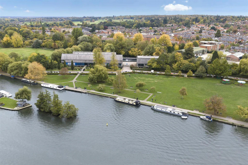 Aerial view of a riverside park with boats moored along the bank, a large modern building, trees with autumn foliage, and a town with houses in the background under a partly cloudy sky.