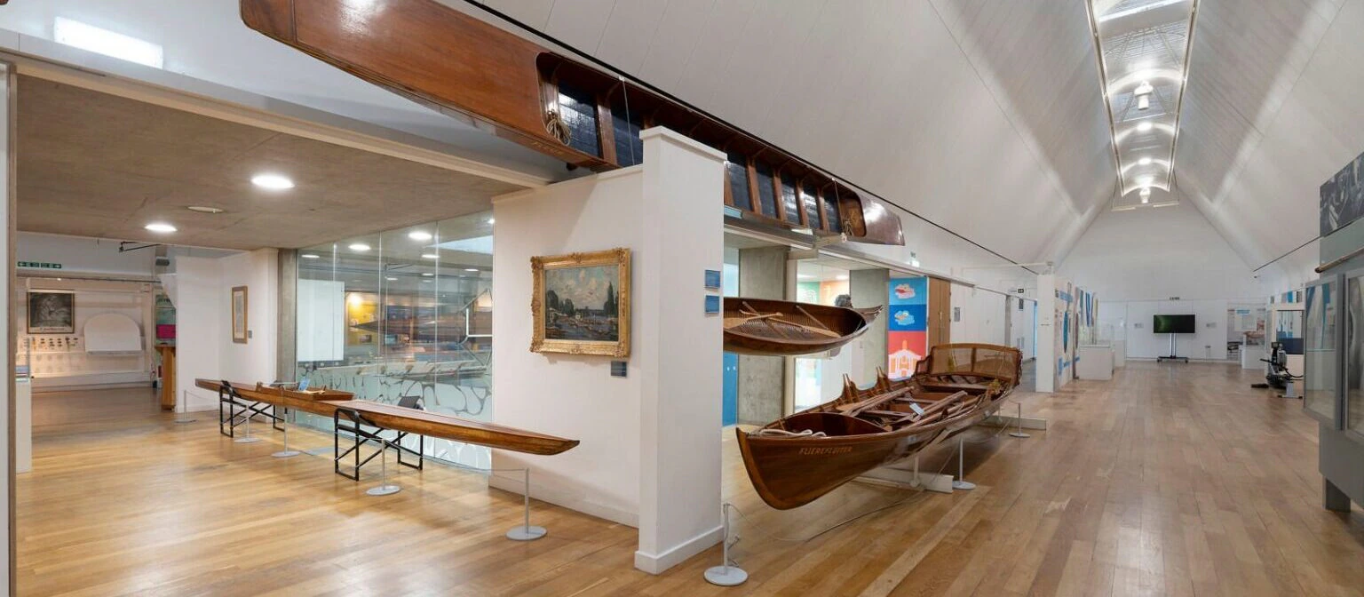 A spacious museum hallway with polished wooden floors displays several wooden boats, paintings on the walls, and informational exhibits. The ceiling is high and arched with skylights, creating a bright, airy atmosphere.