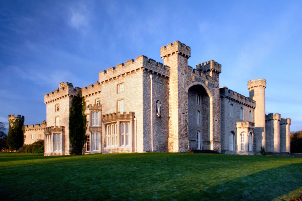 A large stone castle with multiple square towers and battlements, set on green grass and lit by the warm light of sunset against a blue sky.