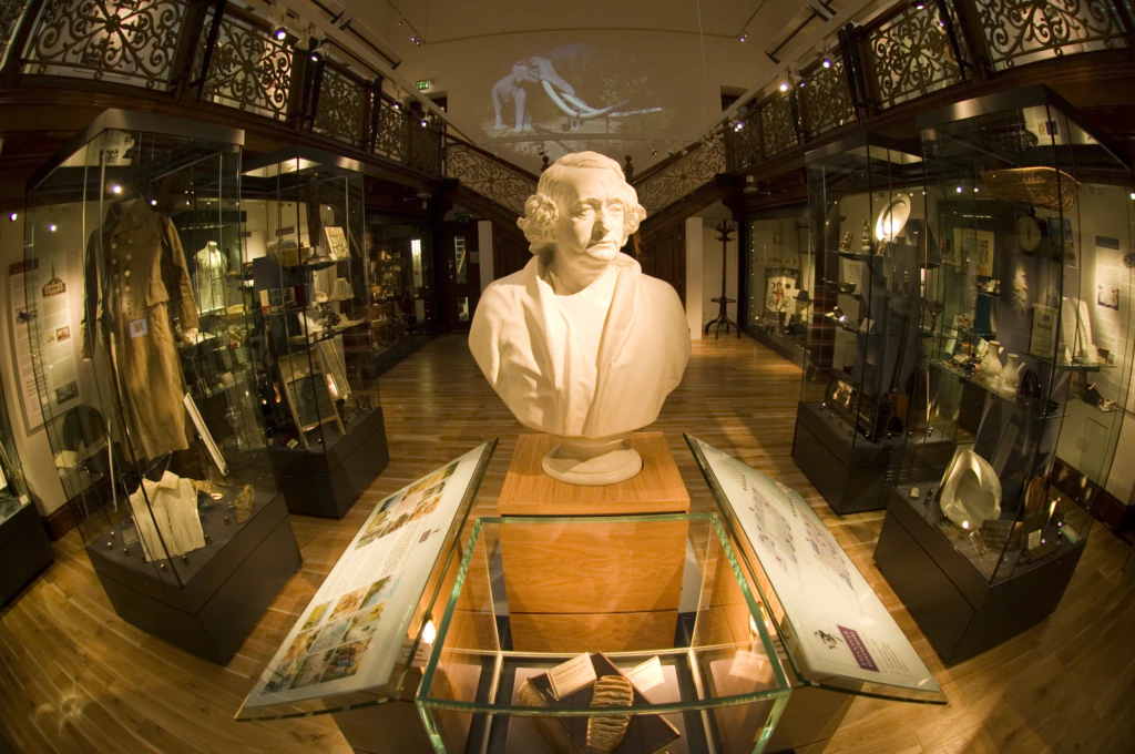 A museum gallery with a central white bust on display, surrounded by glass cases filled with artifacts and historical objects, wooden floors, and ornate railings. A projected image appears on the back wall above the exhibits.