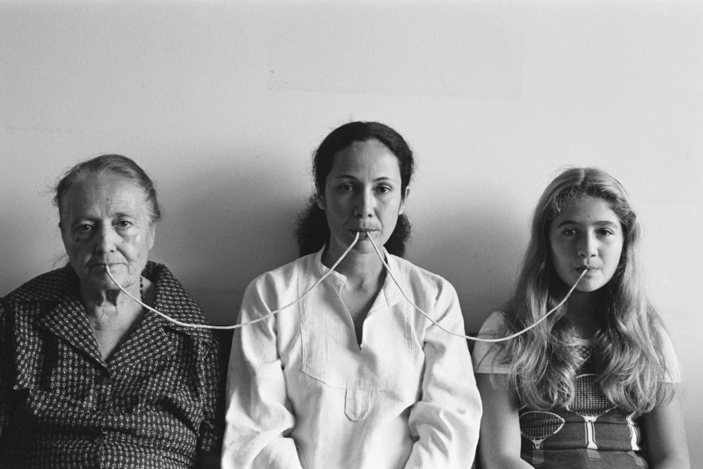 Three women of different ages sit side by side against a plain wall, connected by thin tubes placed in their mouths, forming a visual link among them. The scene appears calm and contemplative.