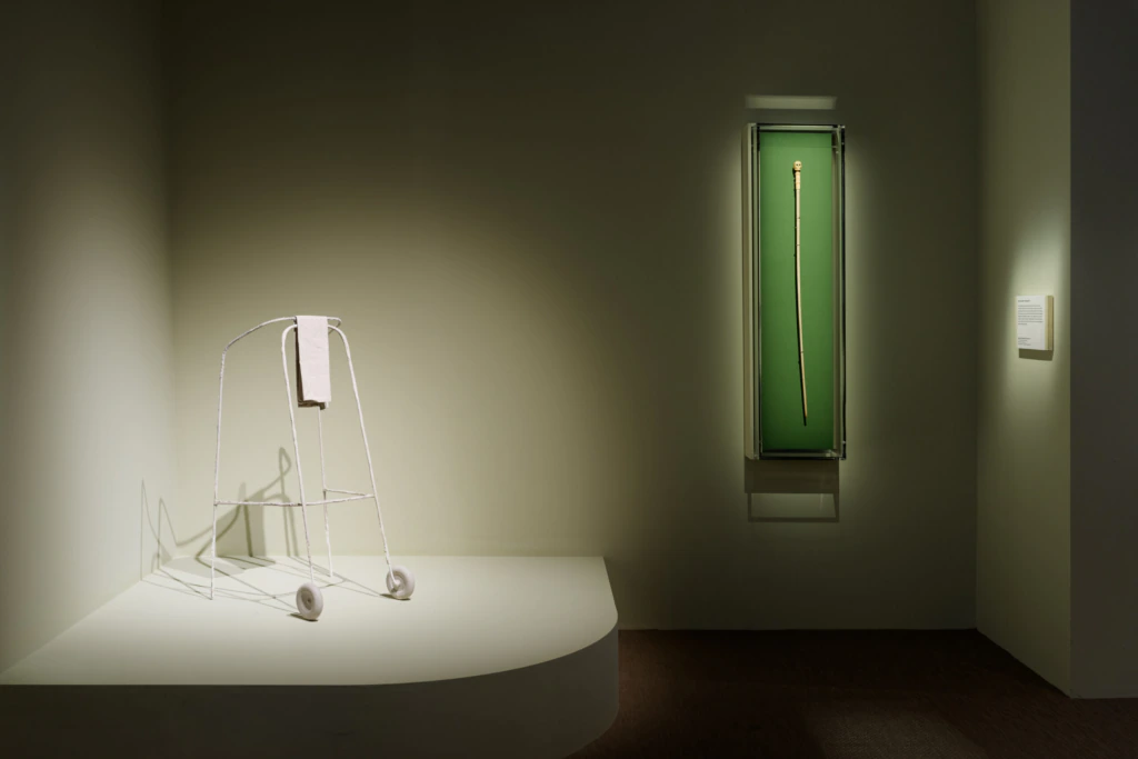 A minimalist display features a white metal walker with a towel draped over it on a platform, and a single cane showcased upright in a green-lit glass case on the wall.