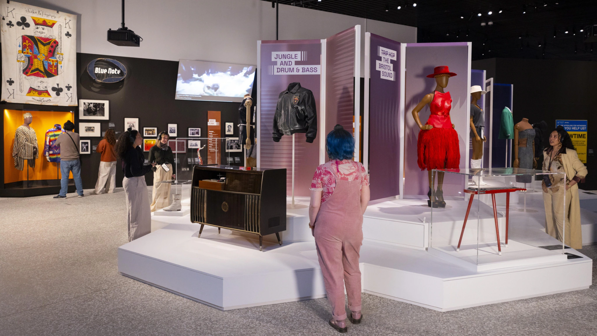 People explore an exhibit featuring vintage clothing, a red dress on a mannequin, a leather jacket, framed photos, and music memorabilia in a modern museum setting.