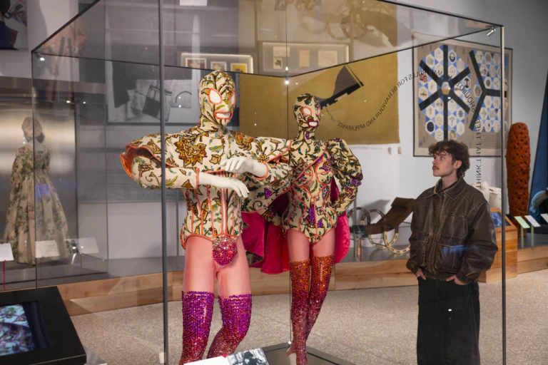 A person observes two mannequins in a museum display case. The mannequins wear colorful, patterned bodysuits with masks and sequined thigh-high boots. The exhibit space includes textiles and artifacts on the walls.