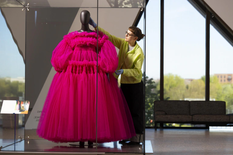 A museum worker in a yellow sweater adjusts a bright pink, ruffled dress displayed on a mannequin inside a glass case. Large windows in the background reveal trees and distant buildings.