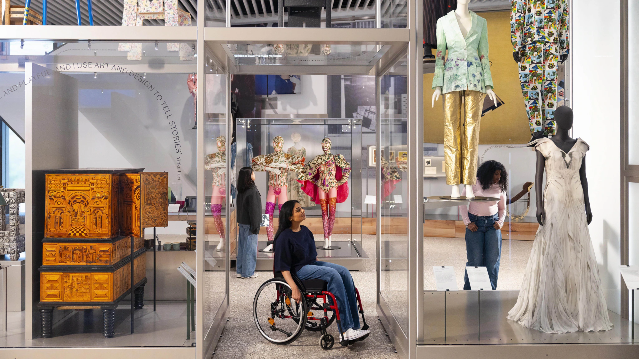 A woman in a wheelchair looks at exhibits in a modern museum, surrounded by colorful costumes and elaborate fashion displays. Another woman in the background views an exhibit while mannequins in unique outfits are showcased nearby.