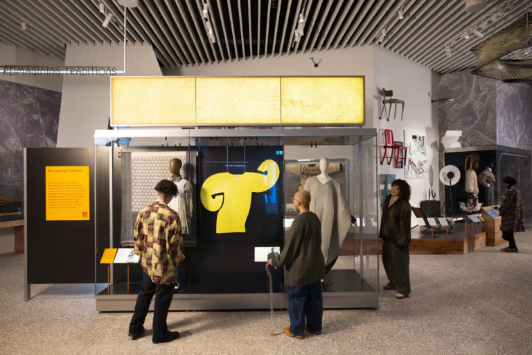 Four people observe a museum exhibit featuring a bright yellow, abstract object displayed in a glass case. The modern gallery has other art pieces and informational panels on the surrounding walls.