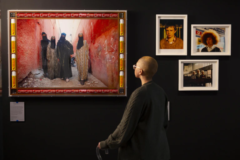 A person stands in an art gallery, viewing a large, framed photo of four veiled women in a red alley. Nearby, three smaller framed portraits hang on a black wall.