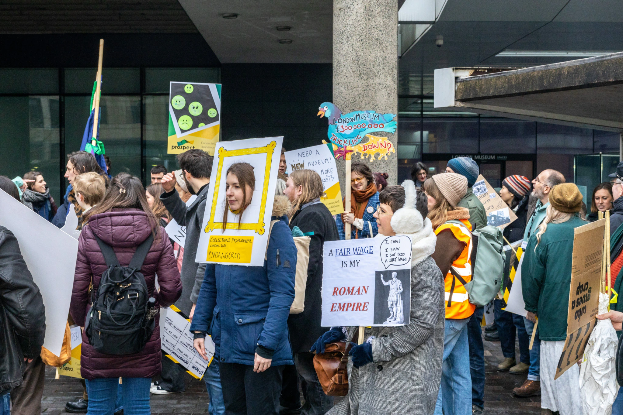 A group of people at a protest, holding signs with messages about fair wages. Some wear yellow safety vests, and the crowd is dressed warmly, suggesting cold weather. One person’s sign is framed like a picture.