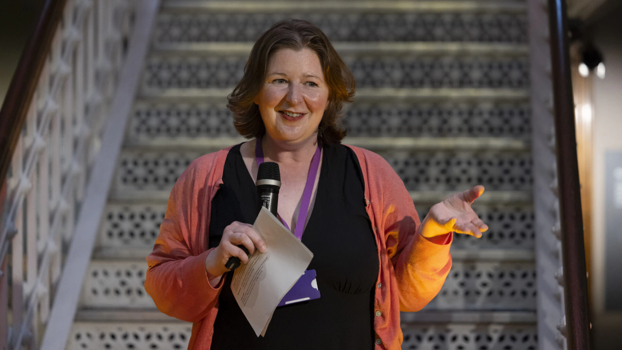 A woman with shoulder-length brown hair, wearing a pink cardigan and black dress, stands in front of a staircase holding a microphone and papers, speaking and gesturing with her hand.