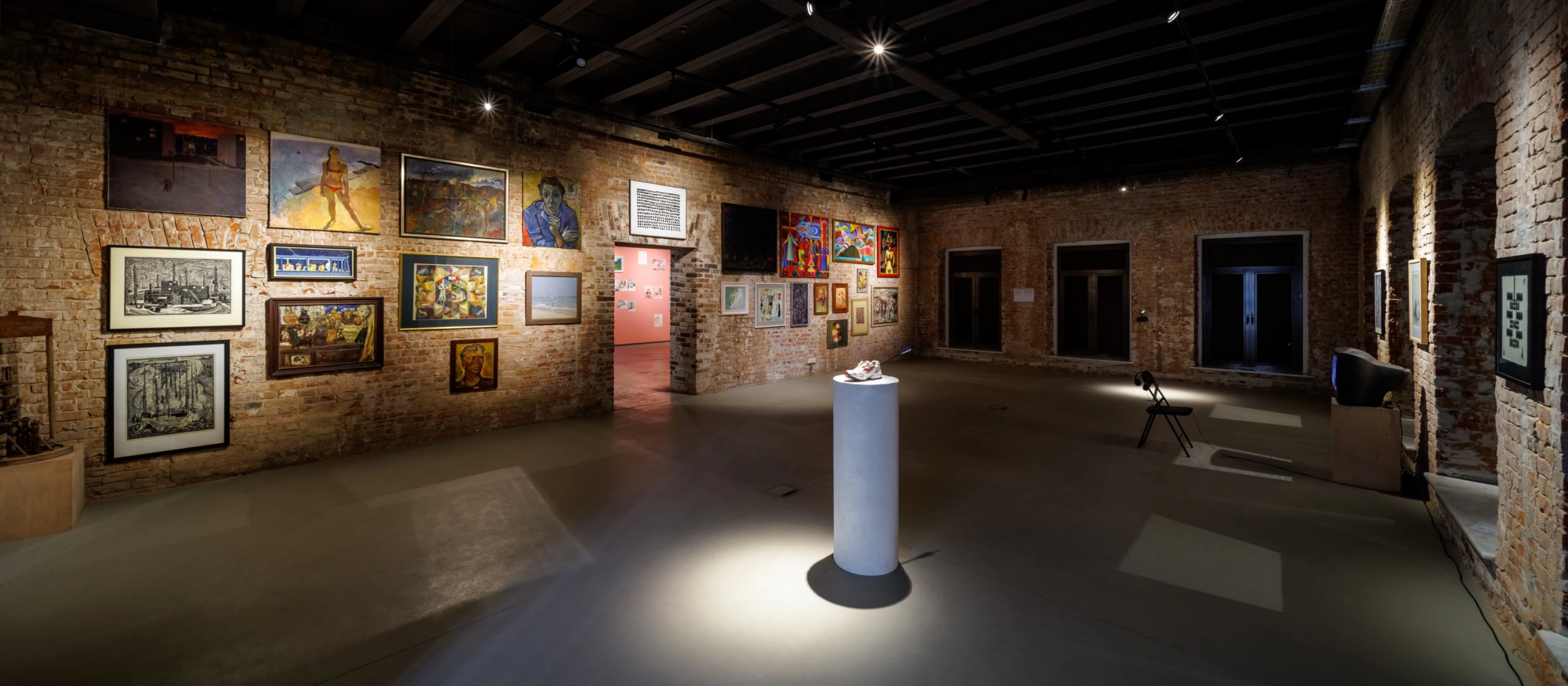 An art gallery with exposed brick walls displays various framed artworks. A single sculpture stands on a pedestal in the center, illuminated by a spotlight. The room is dimly lit with an industrial ceiling and concrete floor.