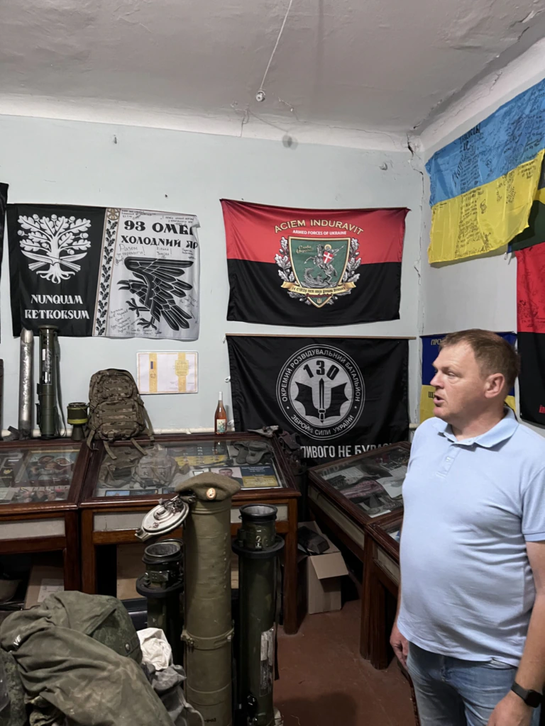 A man in a light blue shirt stands in a room with glass display cases holding military items. Behind him are military flags and a Ukrainian flag hanging on the walls.