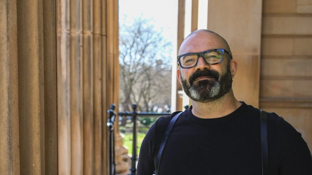 A bearded man wearing glasses and a black shirt with suspenders stands smiling near large stone columns, with trees and sunlight visible in the background.