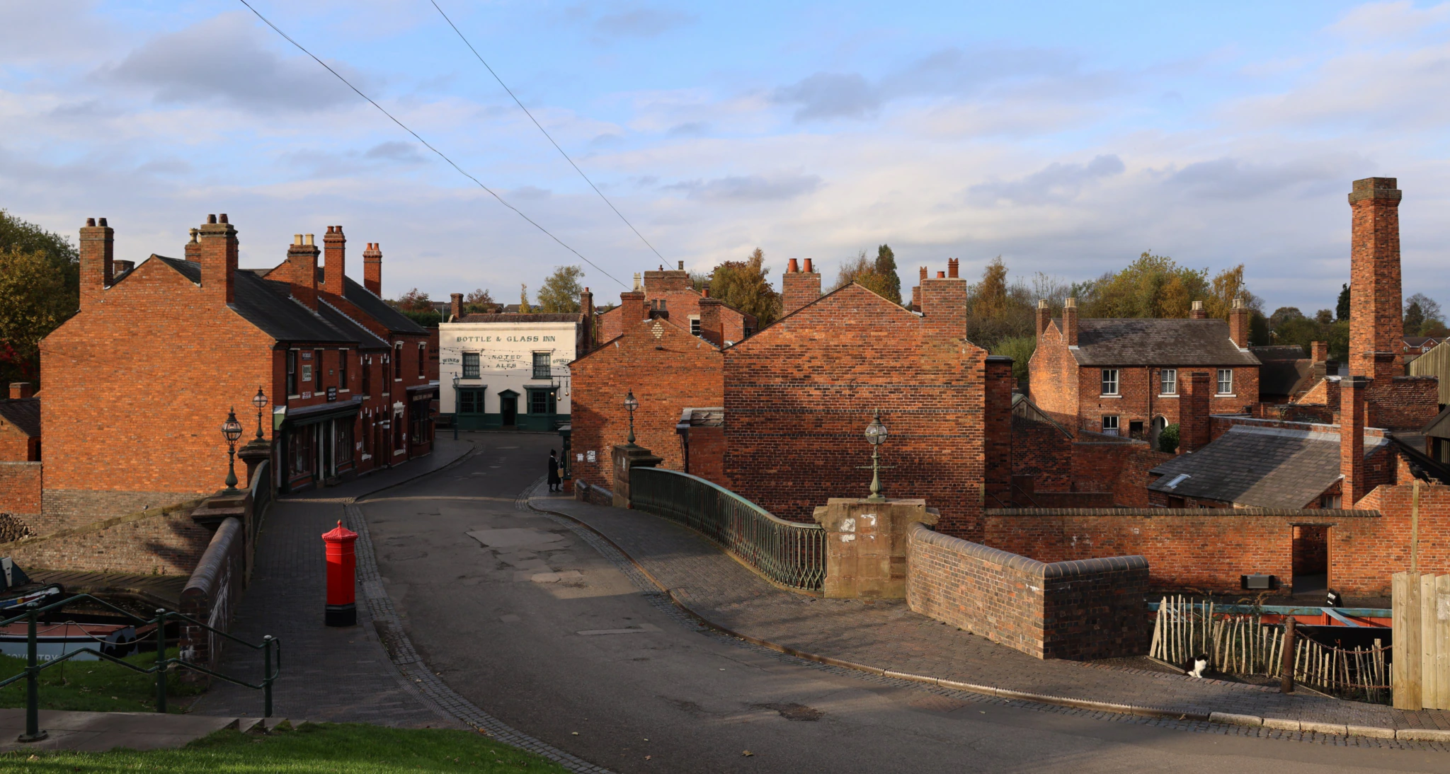 A quiet street in a historic village with red brick buildings, a stone bridge, a red postbox, and a pale blue sky with scattered clouds. Leafy trees are visible in the background.
