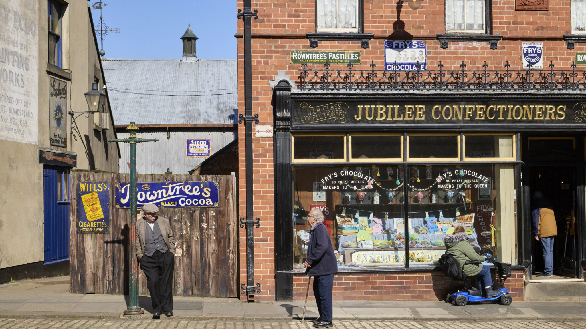 A street scene outside a vintage sweet shop called Jubilee Confectioners. Two men stand talking by a lamppost, while a person on a mobility scooter looks at sweets displayed in the shop window.