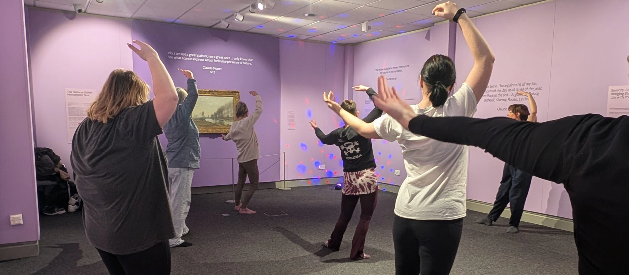 A group of people in casual clothing stand in a circle with arms raised, participating in a movement or dance activity inside a purple-walled room with text and an artwork displayed on the walls.