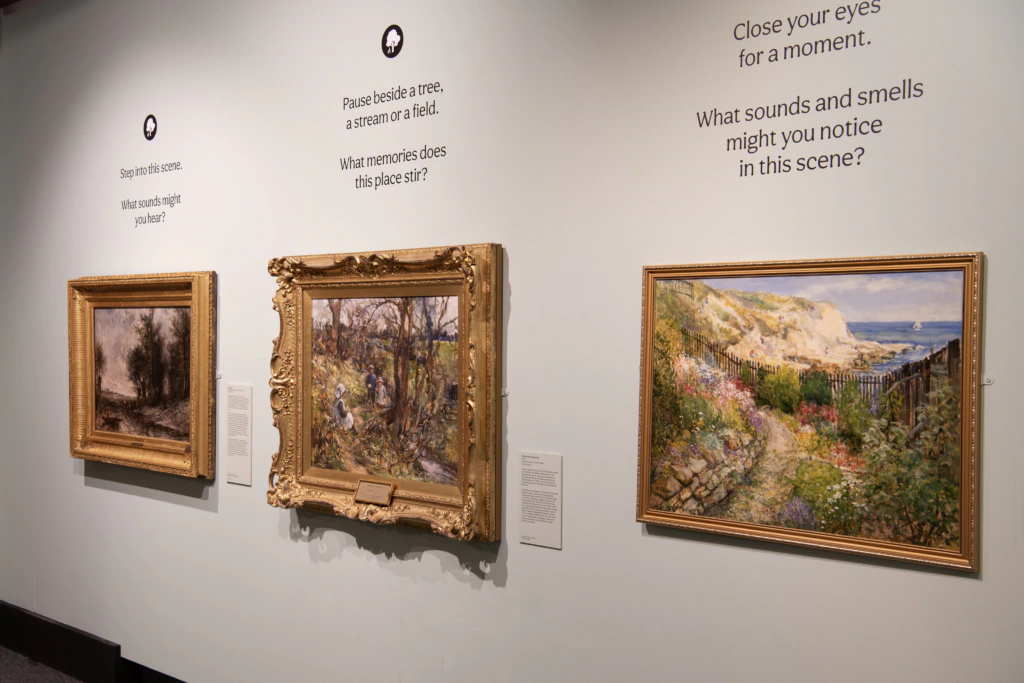 Three framed landscape paintings hang on a museum wall, accompanied by prompts encouraging viewers to reflect on the scenes, memories, and senses evoked by the artwork.