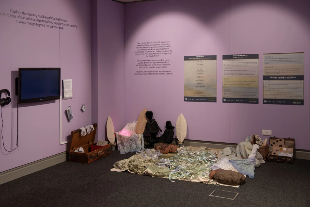 A museum exhibit room with pastel purple walls displays an installation of fabric and soft sculptures on the floor, a wall-mounted TV screen with headphones, and several informational posters on the wall.