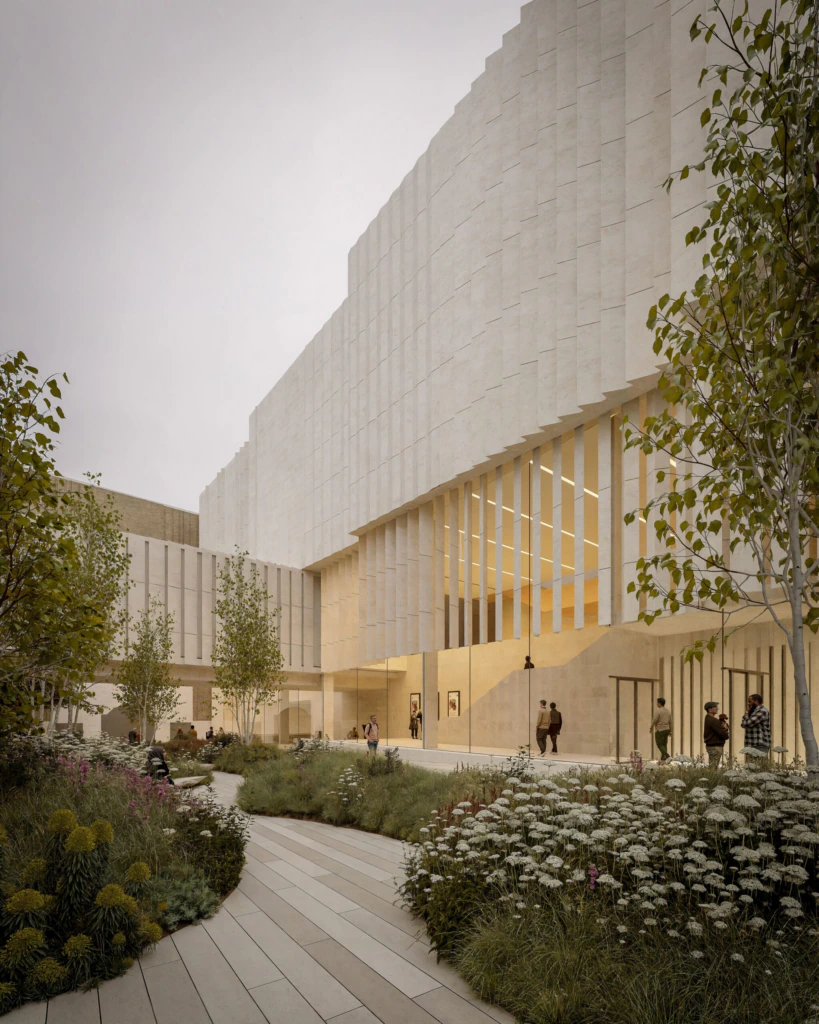 A modern building with textured light stone walls and tall vertical windows faces a landscaped courtyard with trees, flowering plants, and people walking or standing on paved paths.