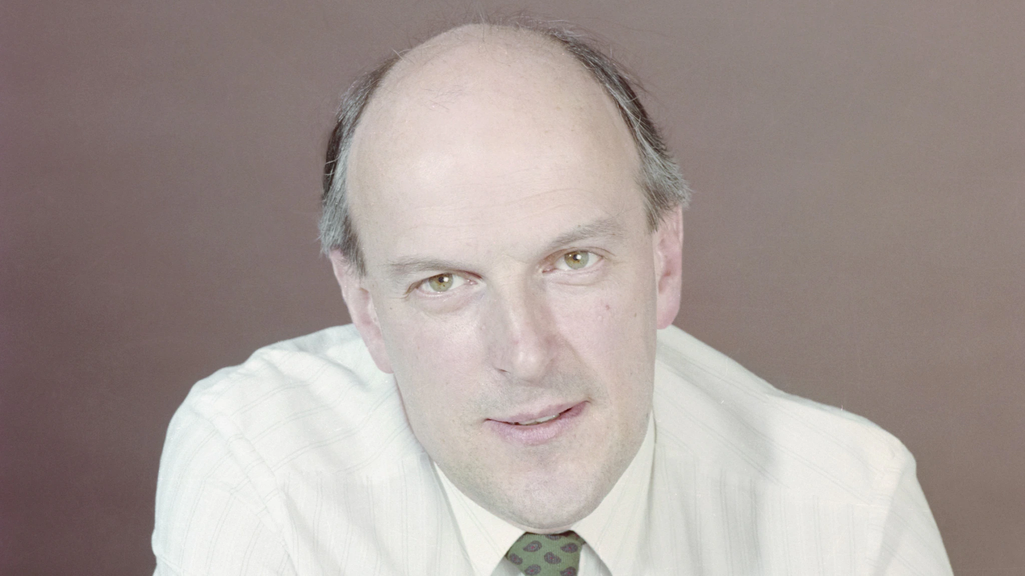 A middle-aged man with a receding hairline wearing a light-striped shirt and a green patterned tie, looking directly at the camera against a plain, muted background.
