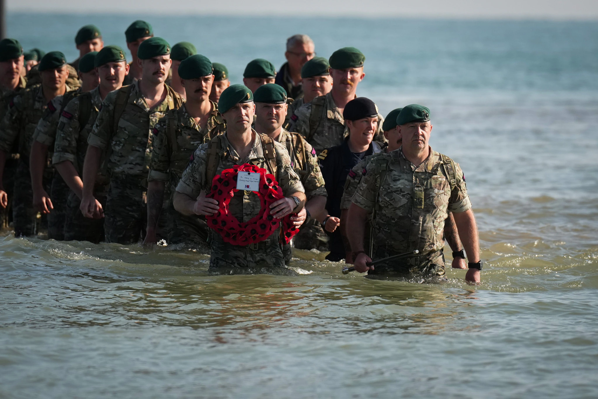 A group of soldiers in camouflage uniforms and green berets wade through shallow water, with one soldier at the front holding a poppy wreath. The background shows a calm sea and a clear sky.
