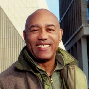 A man wearing a green jacket smiles while standing in front of the V&A Museum. The building has a modern, geometric facade and large V&A letters on top. The image has film borders and light leaks.