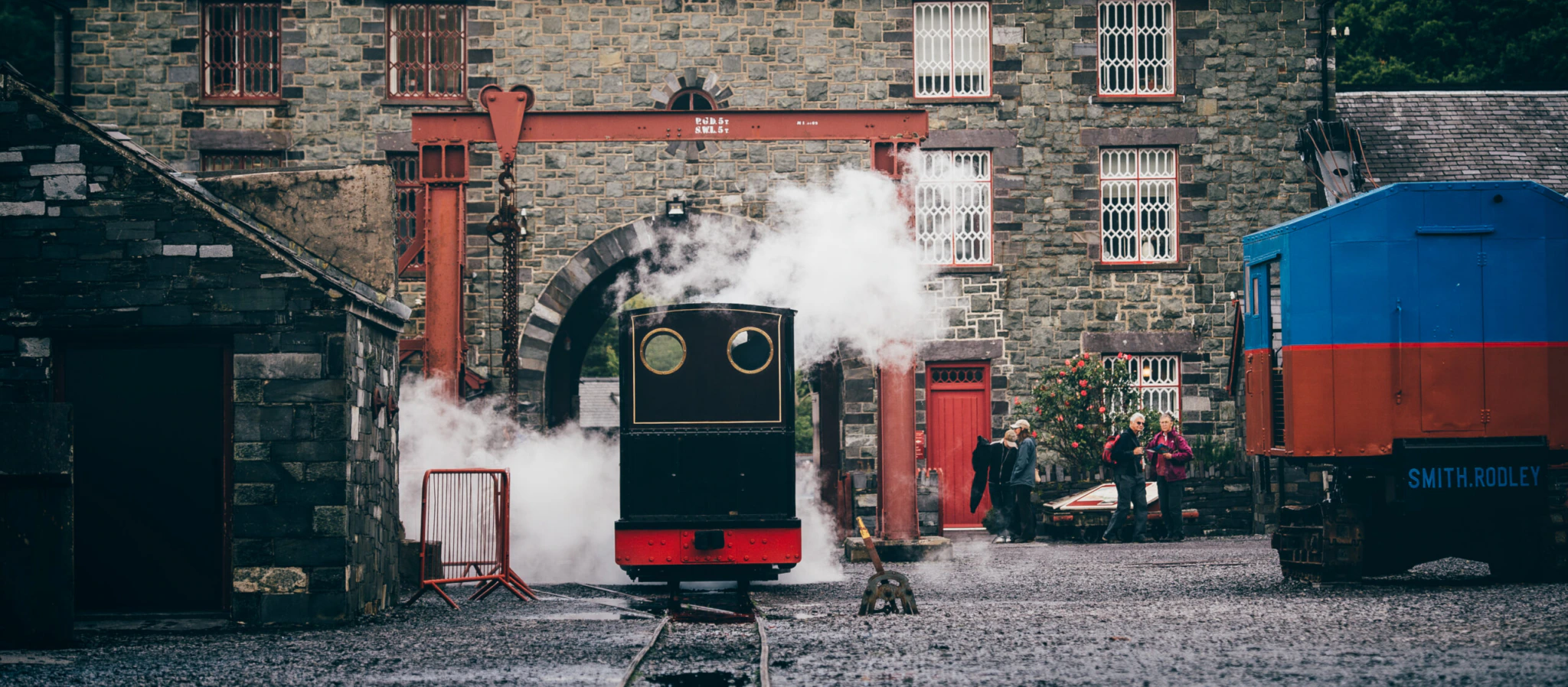 A small steam train with a black and red front releases steam on tracks in front of a historic stone building with red window frames. People stand nearby, and a blue train car is on the right. Lush green trees are in the background.