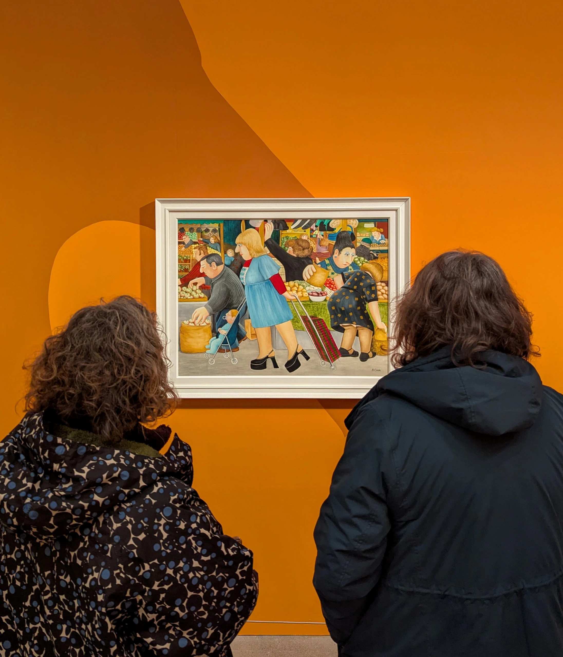 Two people in dark coats stand in front of an orange wall, viewing a framed painting that depicts a lively market scene with several figures engaged in various activities.