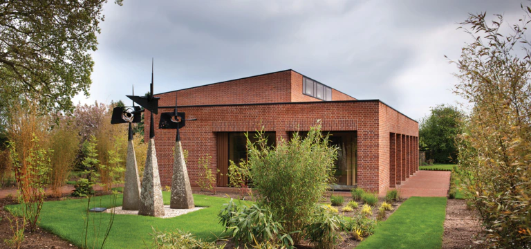 A modern brick building with large windows is surrounded by neatly landscaped gardens. In the foreground, there is a metal abstract sculpture with three tall angled bases and geometric shapes on top.