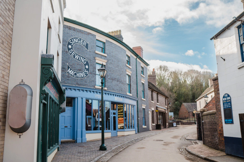 A cobblestone street lined with historic brick buildings, including a blue-fronted shop with a large Singer Sewing Machines sign, under a cloudy sky in a heritage museum setting.