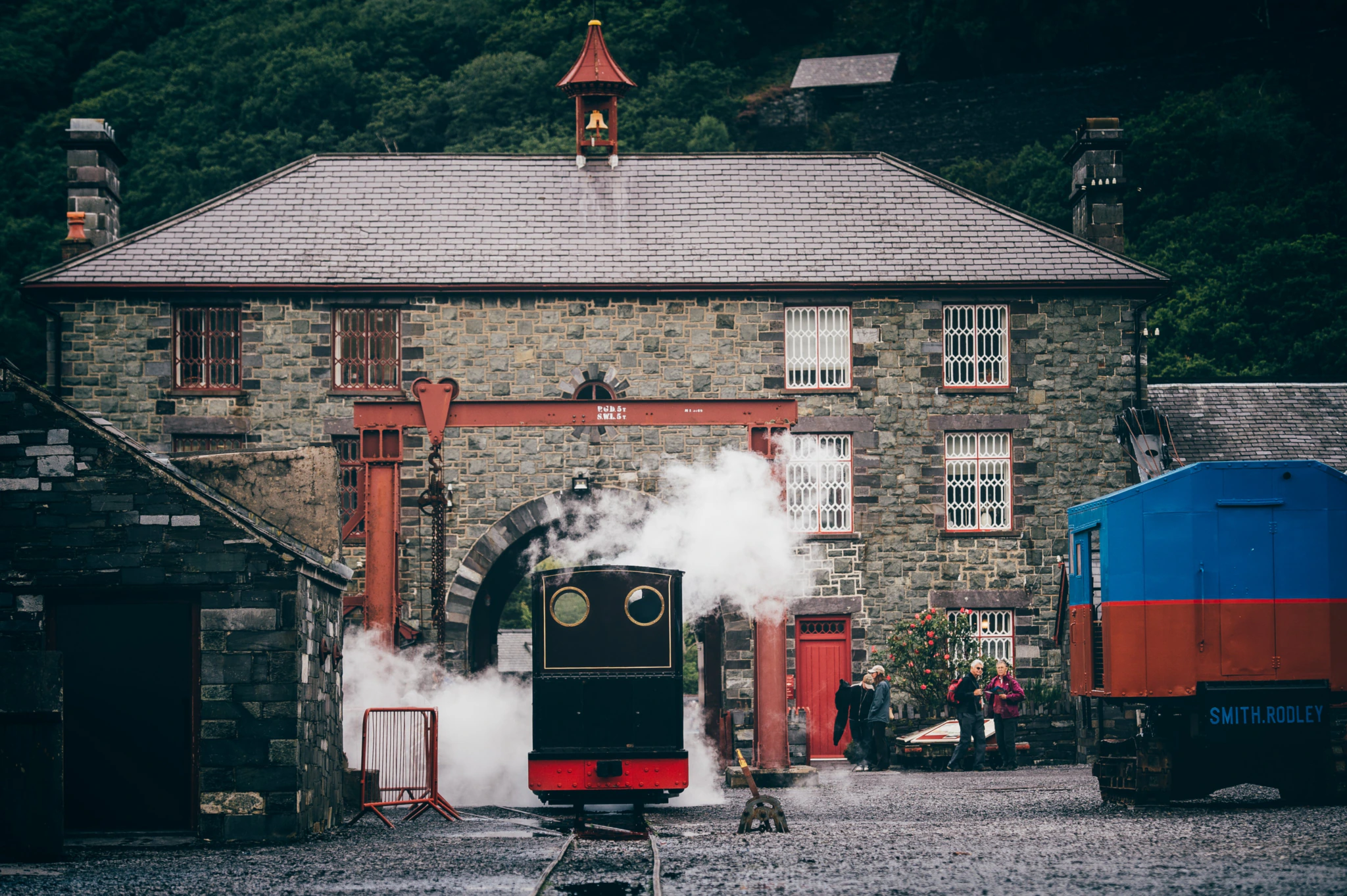 A small black steam train with red trim emits steam in front of a stone building with arched windows and a clock tower. People stand nearby, and a blue rail car is on the right. Lush green trees fill the background.